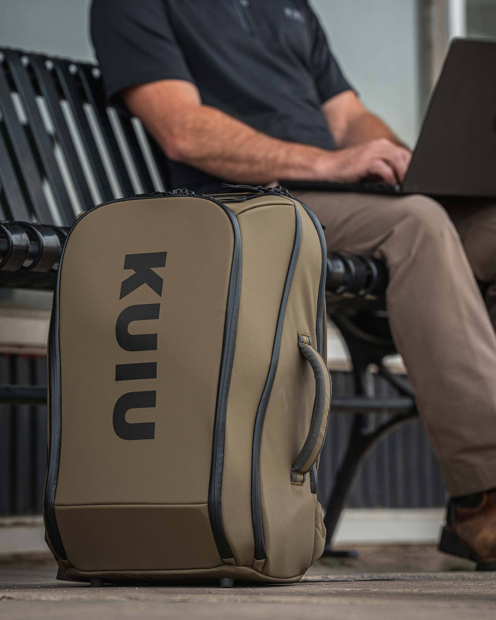 Waypoint Travel Backpack placed on the ground next to a person sitting on a bench using a laptop, highlighting the bag's shape, handle and zipper construction.