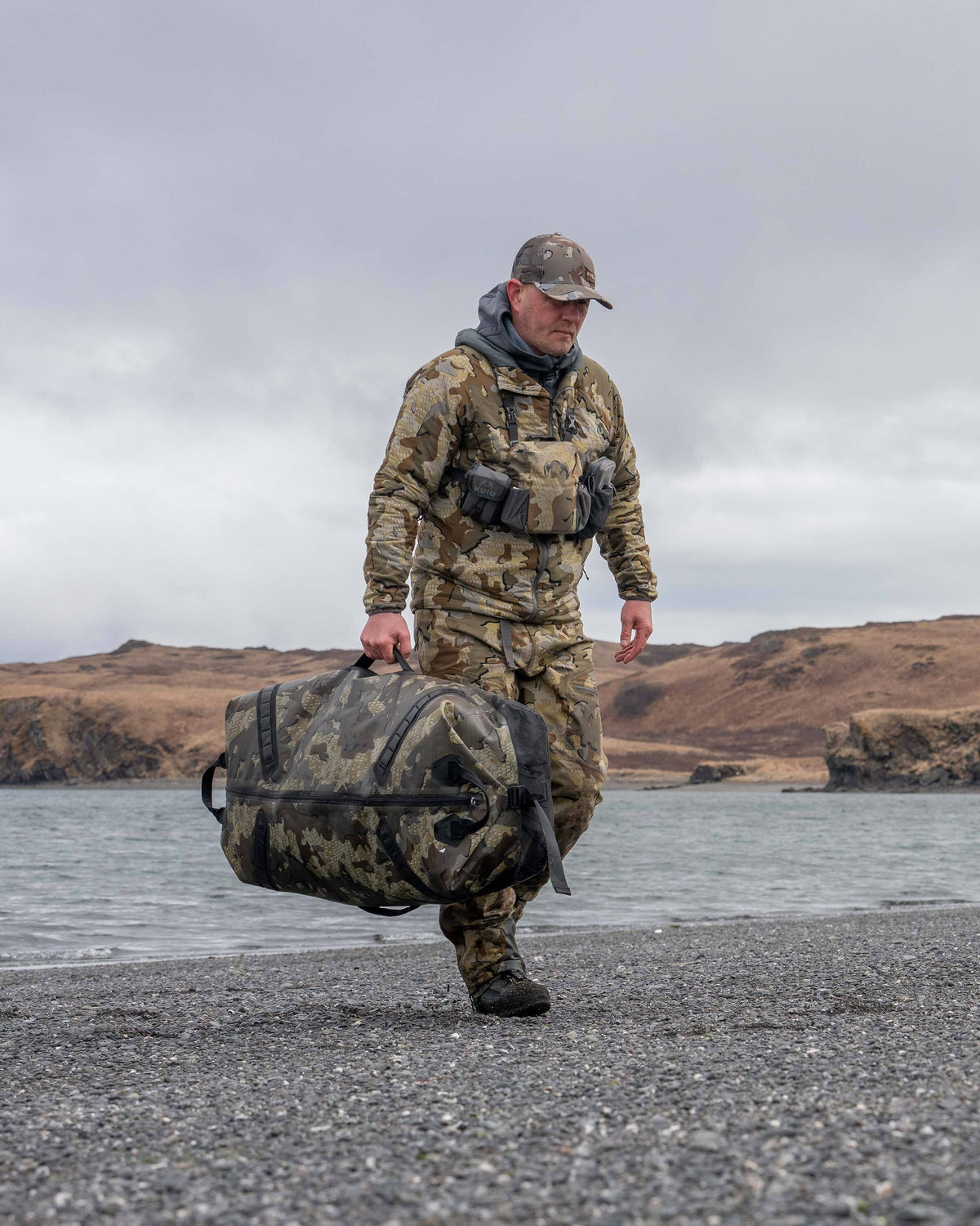 Hunter Walking on Rocky Beach Utilizing Durable Carry Handle on Kodiak 6600 Submersible Duffel in Valo Camouflage