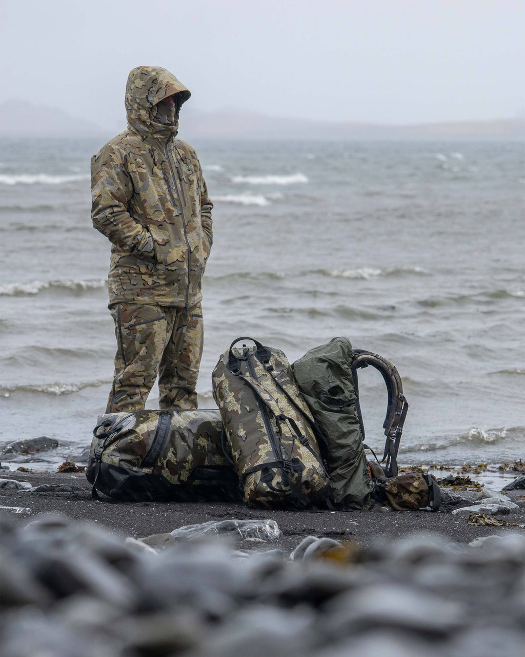 Hunter Standing on Rocky Beach with White Capped Waves in the Background with Kodiak 6600, Kodiak 3000 and Pro Pack in Valo Camouflage
