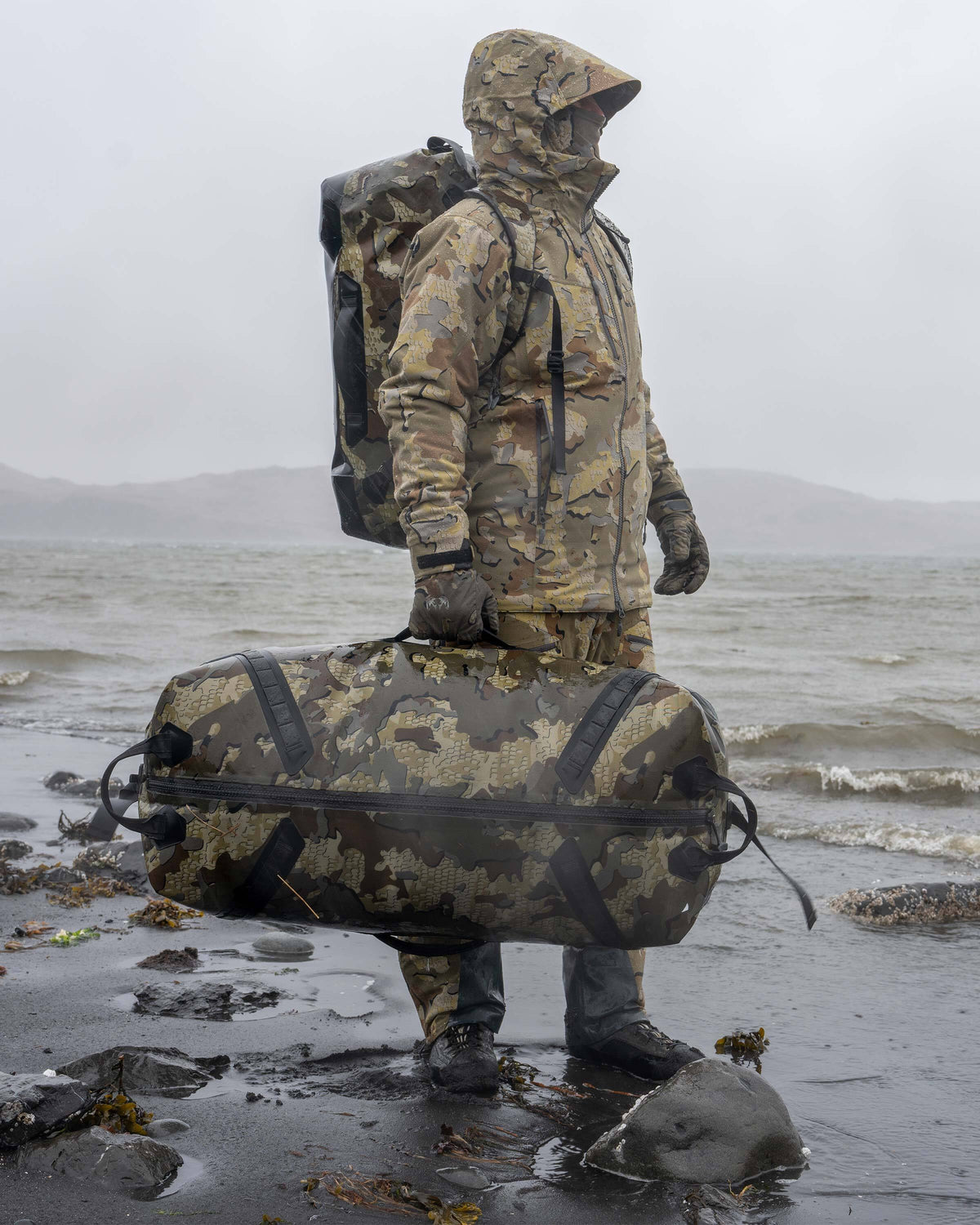 Hunter Standing on Foggy Beach Holding Kodiak 6600 Submersible Duffel in Valo Camouflage
