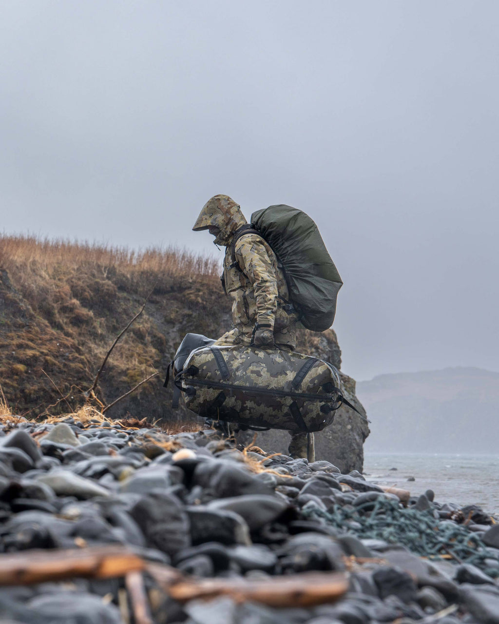 Hunter Walking up Rocky Beach Carrying Kodiak 6600 Submersible Duffel in Valo Camouflage