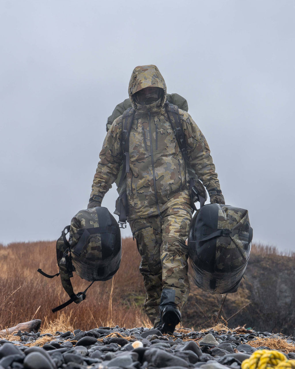 Front of Hunter Walking on Rocky Beach Carrying Kodiak Submersible Duffels in Valo Camouflage Highlighting 840D High Tenacity Hypalon bottom panel