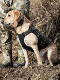 A retriever wearing a black and Valo camouflage neoprene dog vest sits outdoors on dry ground next to a person in Valo camouflage clothing.