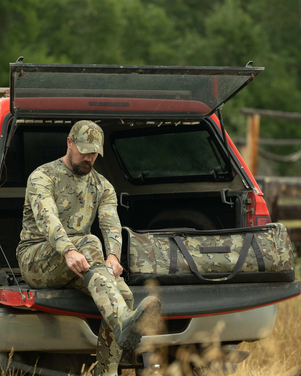 Hunter seated on the tailgate of his truck, is putting on his boots while next to the TwoTrack 8000 Boot Duffel in Valo Camouflage