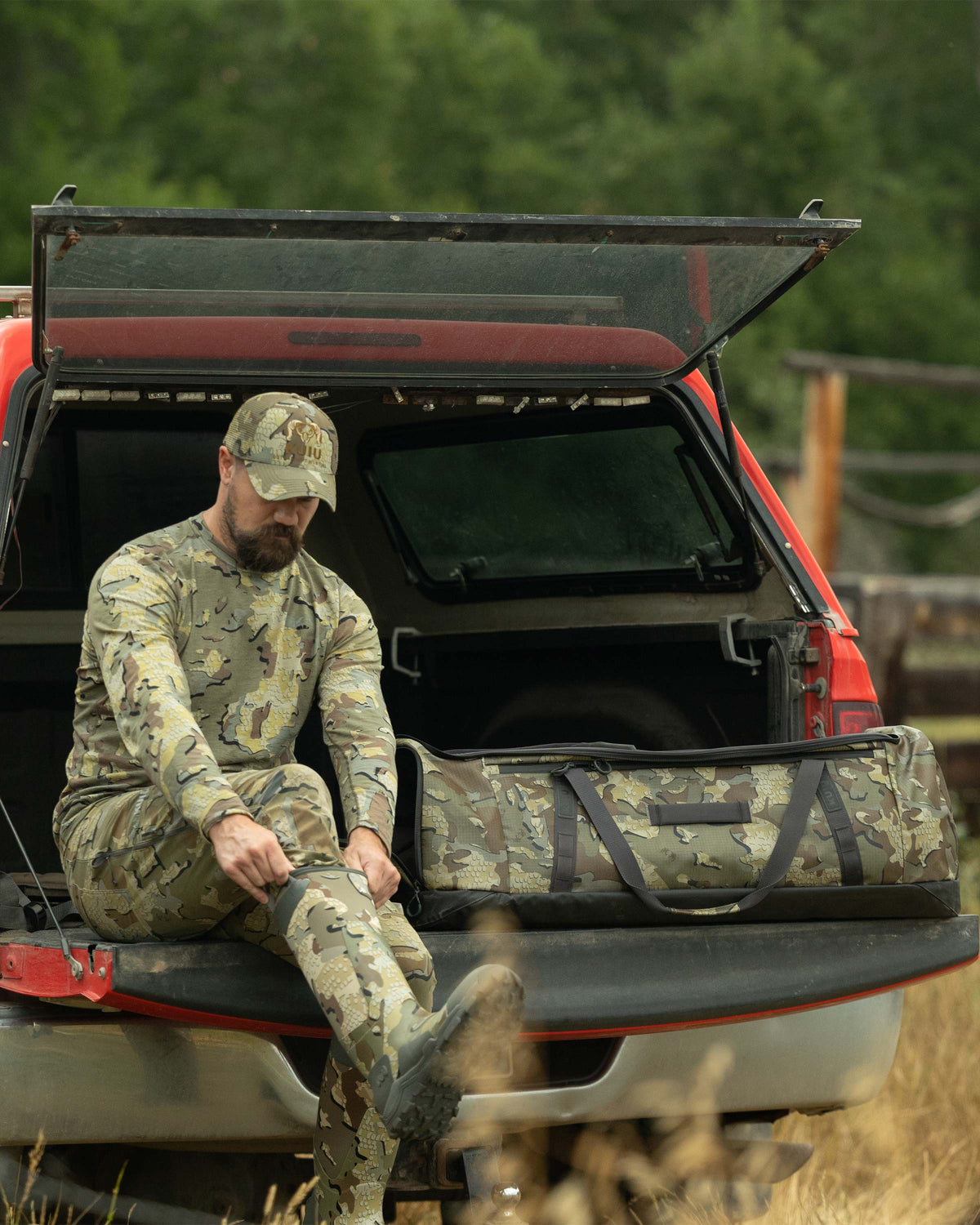 Hunter seated on the tailgate of his truck, is putting on his boots while next to the TwoTrack 8000 Boot Duffel in Valo Camouflage