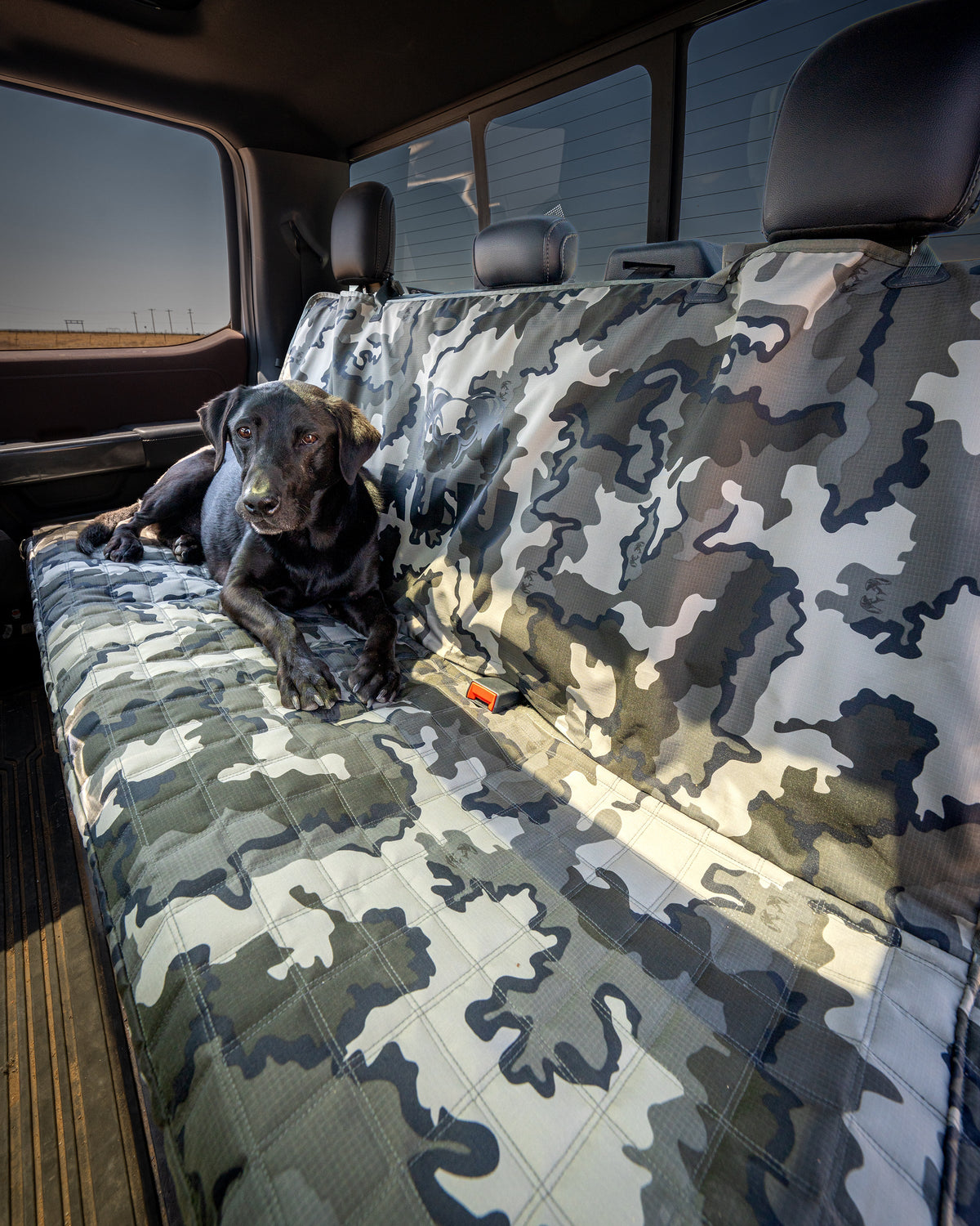 A black dog lies on a large backseat cover with the Quilted K9 Seat Cover in Vias Storm inside a vehicle. The cover is quilted and covers the entire back seat area.