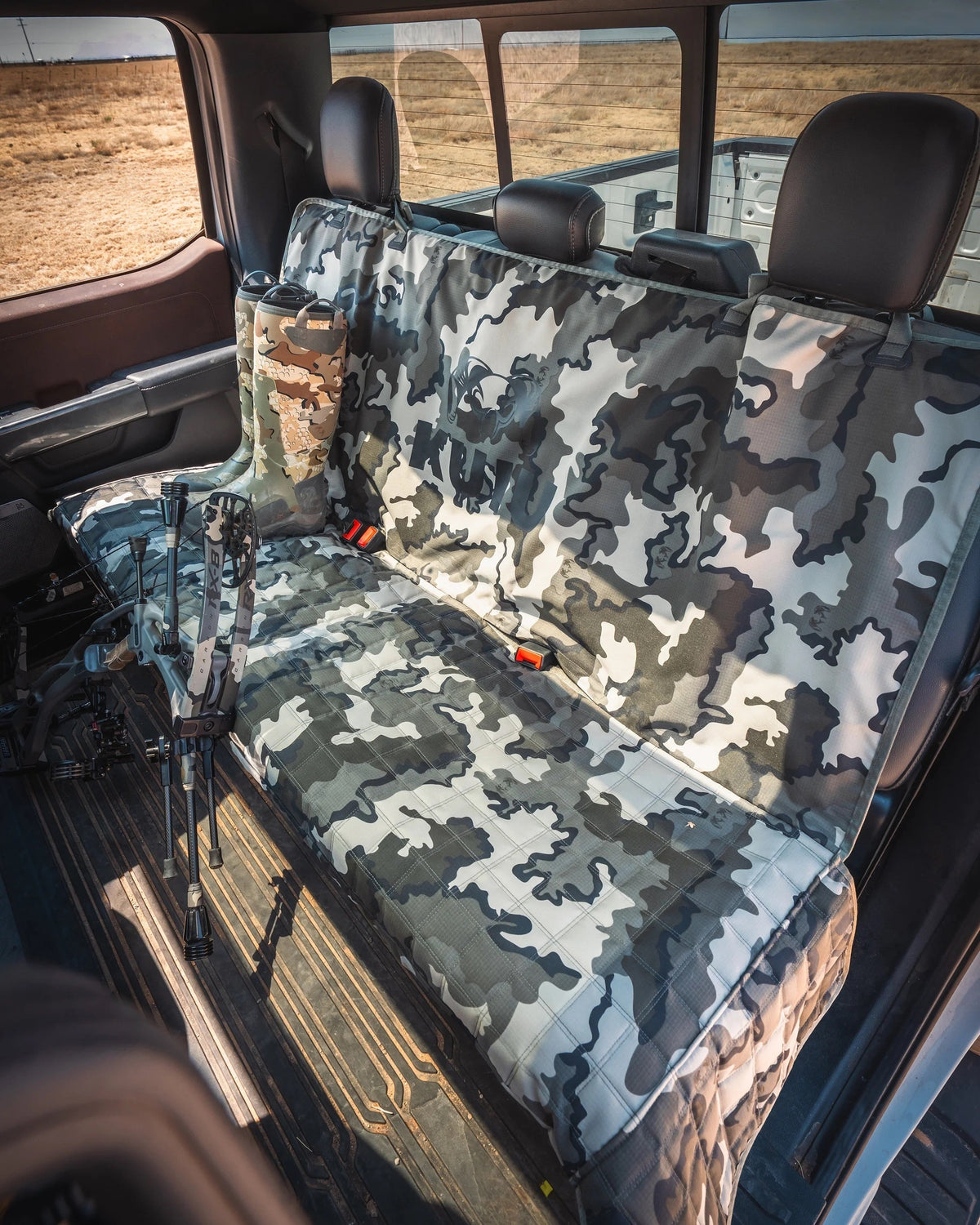 KUIU Quilted K9 Seat Cover in Vias Storm camouflage seat covers installed on the rear bench of a vehicle, with a tripod visible on the floor and an outdoor landscape visible through the windows.