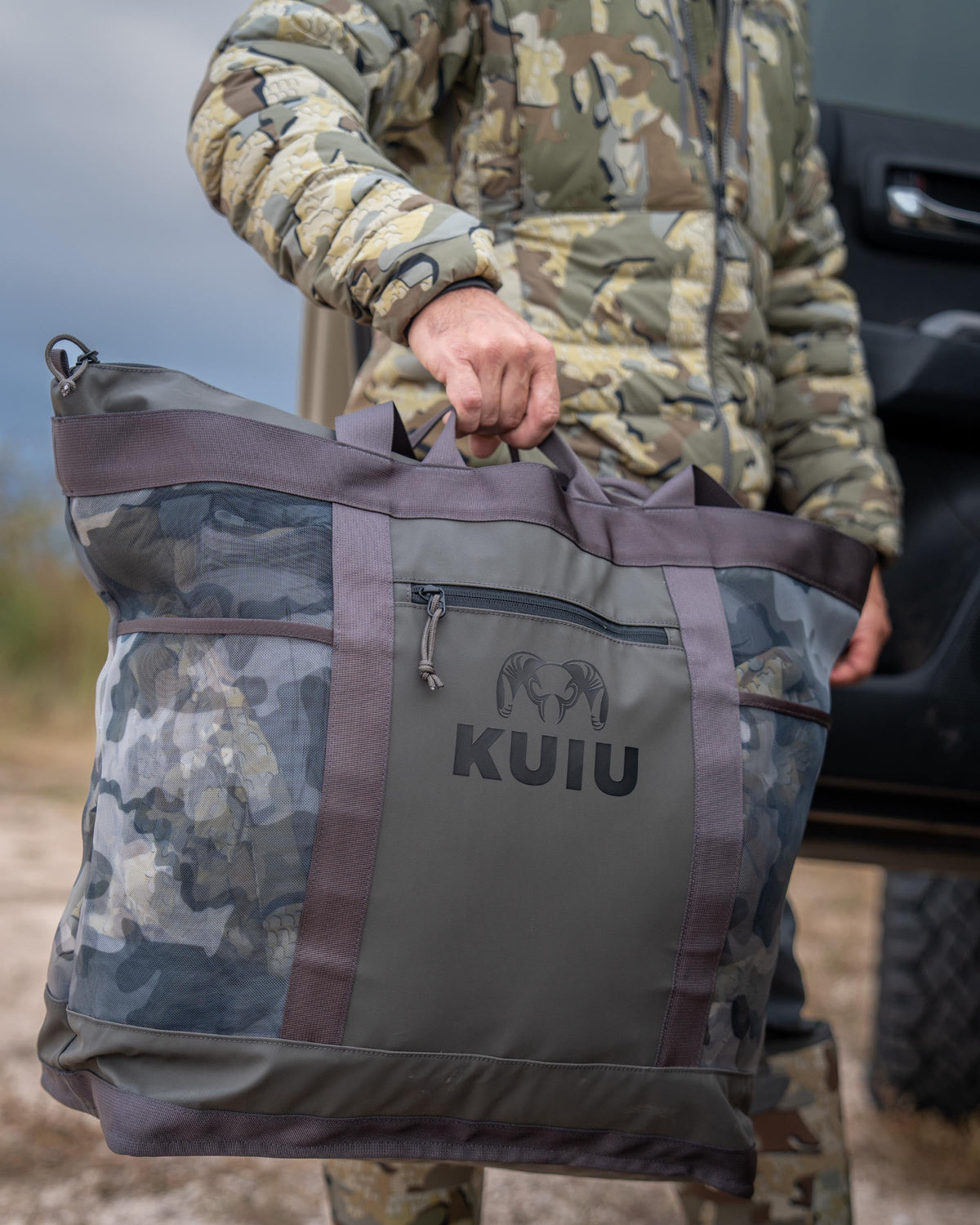 Hunter holds up KUIU 3500 Mesh Tote Bag in Vias Storm, Highlighting front material and zippered pocket