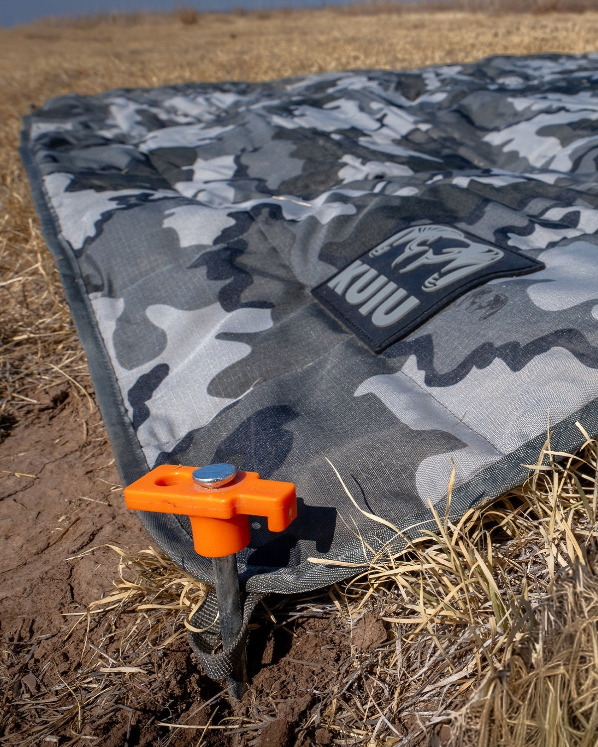 KUIU Distance AT Blanket laid out in an outdoor field, secured with an orange ground anchor. 