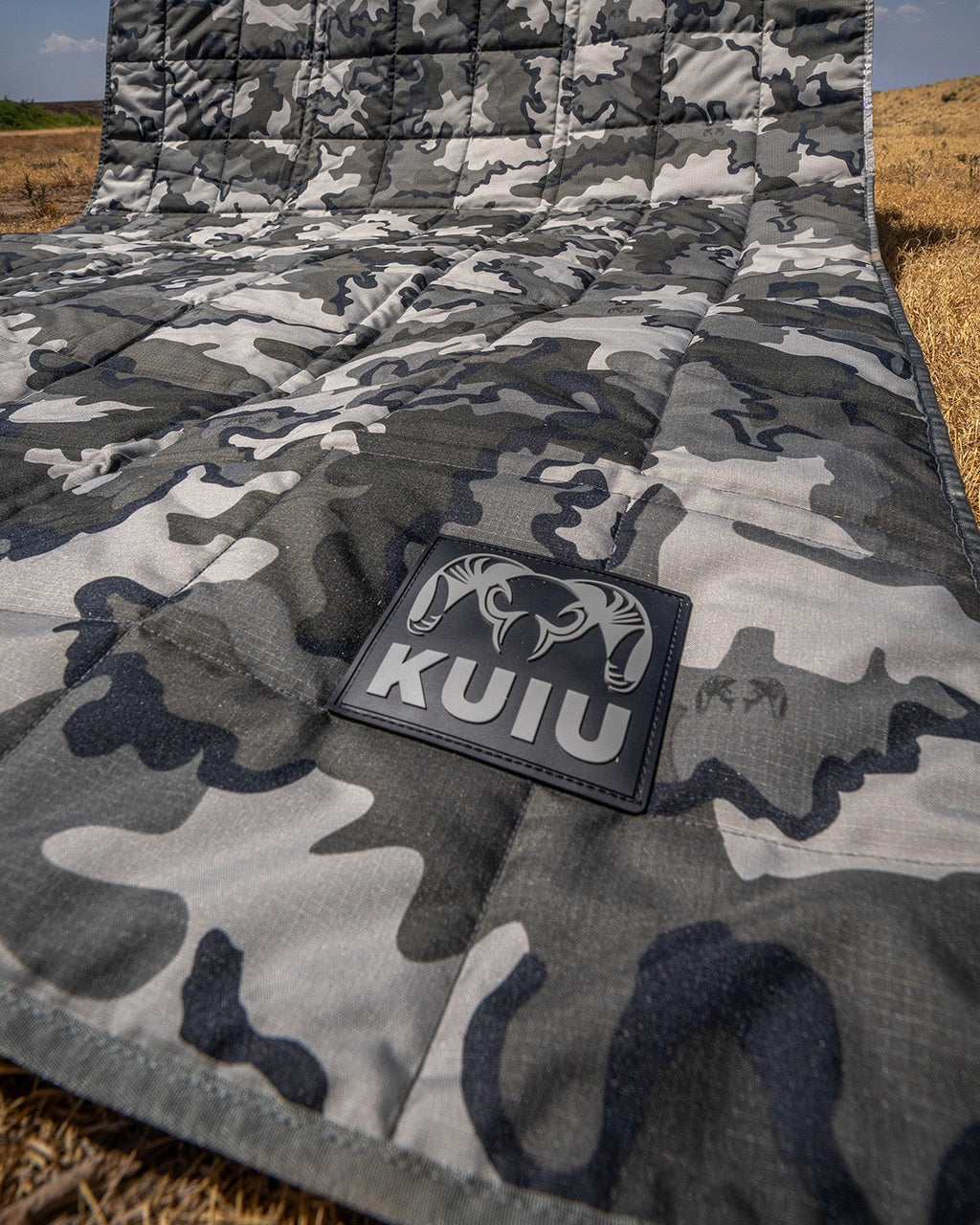 Distance AT Blanket shown with a sewn-in black rectangular patch featuring a ram logo and the brand name 'KUIU', laid out outdoors on dry grass.