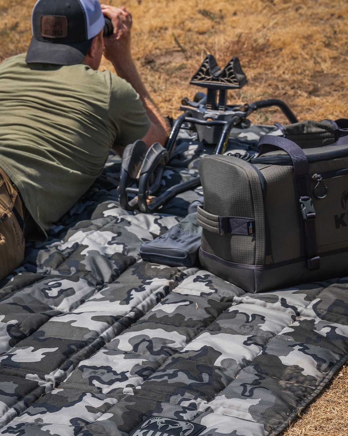 A man lies on a Distance AT Blanket in Vias Storm outdoors on dry grass, looking through a binocular with a dark gray carrying case and a tripod visible nearby.