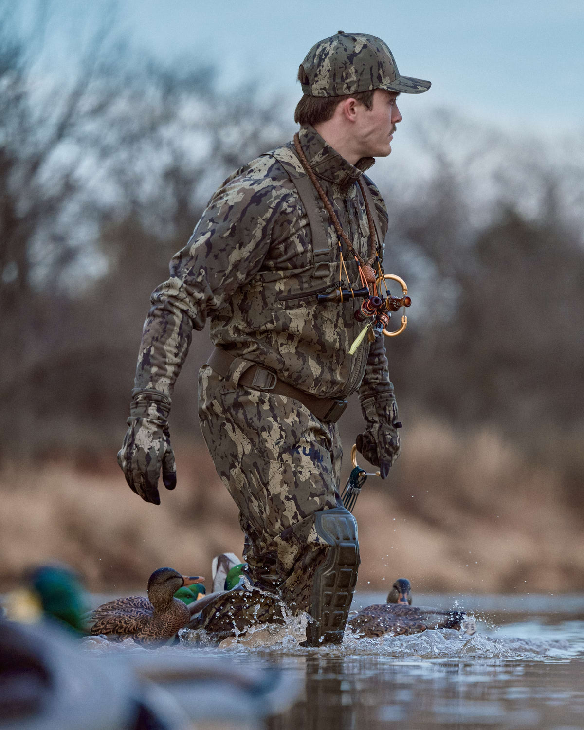 Waterfowl hunter wearing StrongFleece 290 Glove in Veros Camouflage stands in shallow water near duck decoys. The background shows a blurred natural outdoor setting with trees and reeds.
