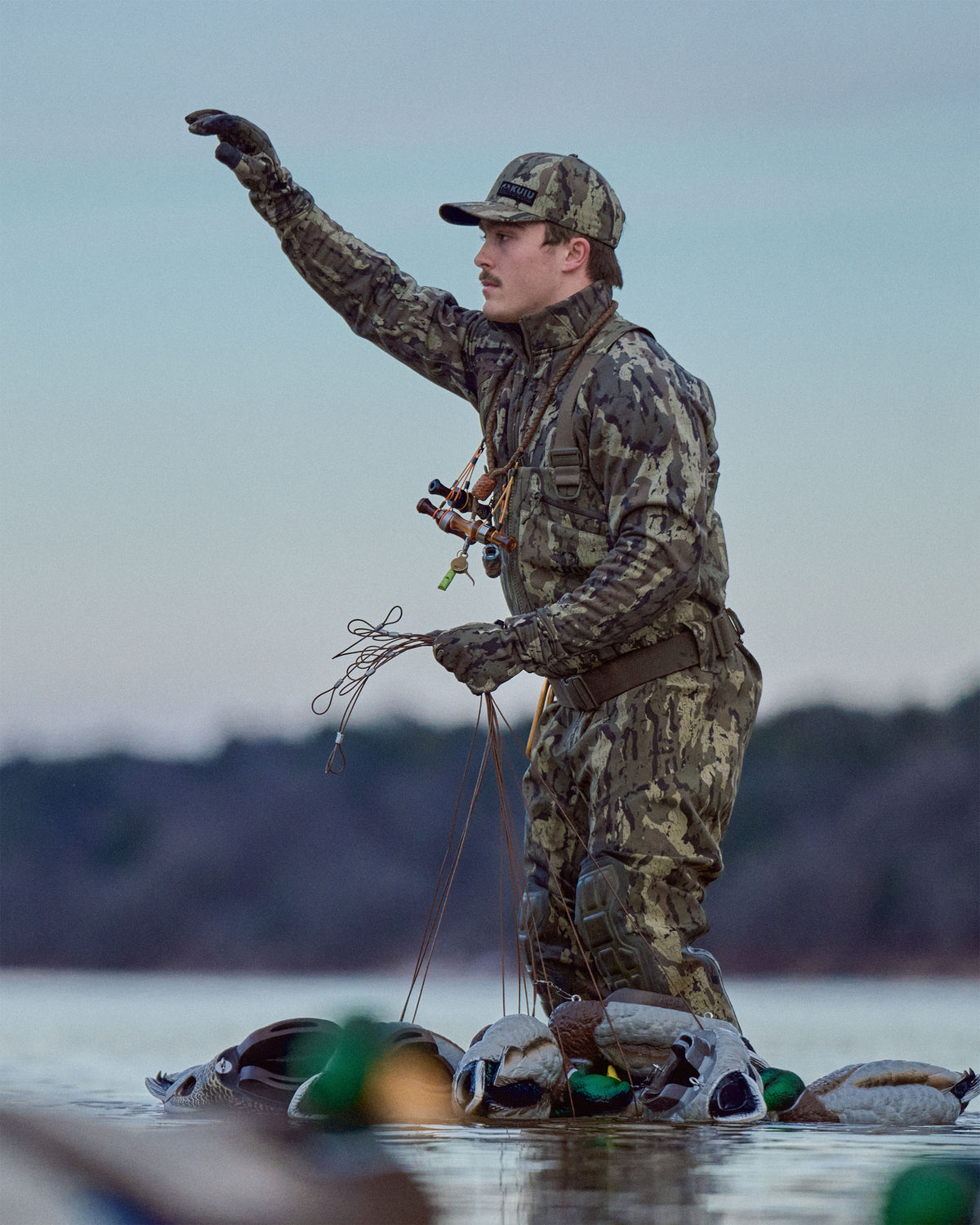 Waterfowl hunter wearing KUIU PRO HD Flex Zip Wader and StrongFleece 290 Gloves in Veros Camouflage stands in shallow water holding half a dozen of duck decoys.