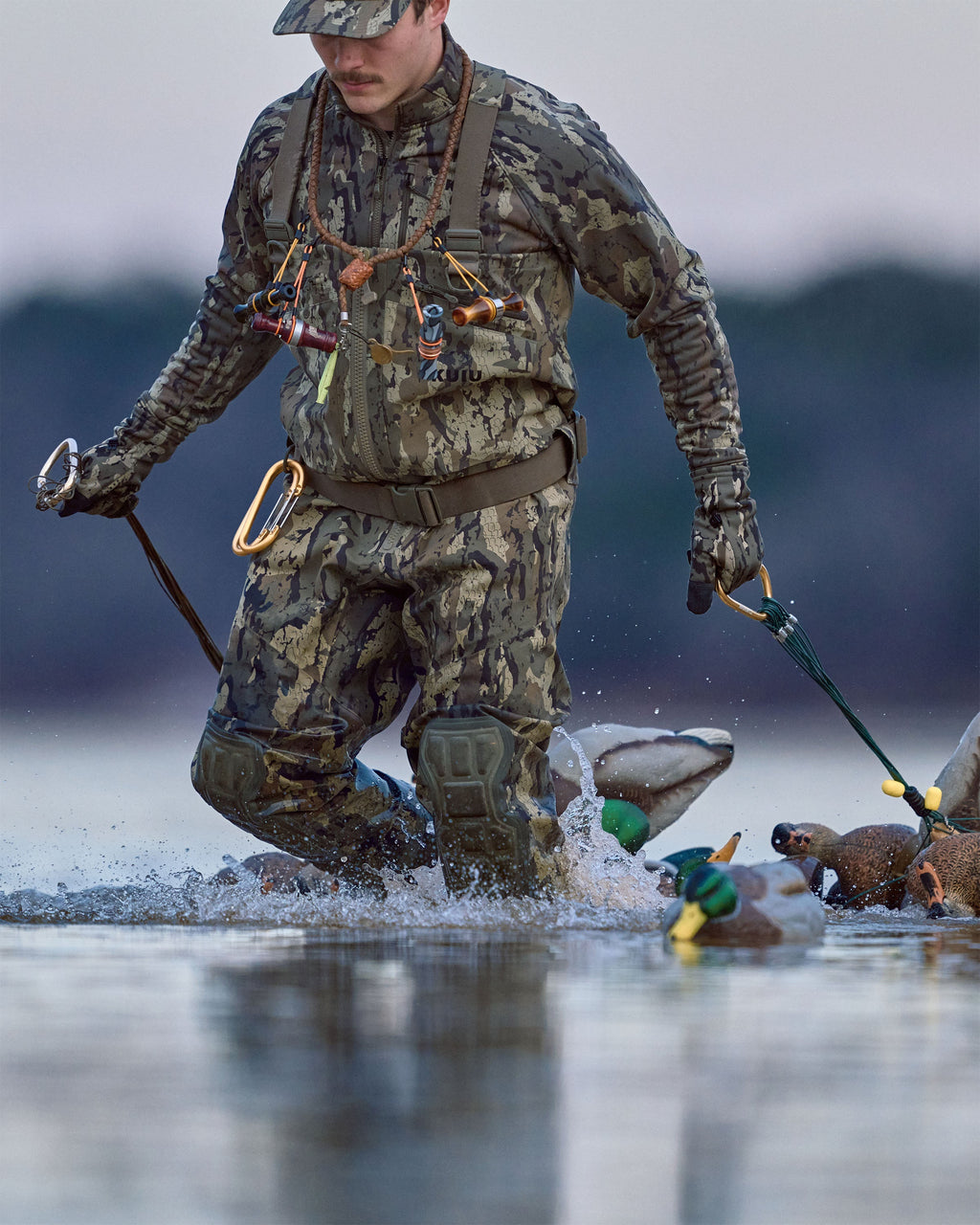 Waterfowl hunter wearing KUIU PRO HD Flex Zip Wader and StrongFleece 290 Gloves in Veros Camouflage wades through water while pulling a floating string of duck decoys behind him.