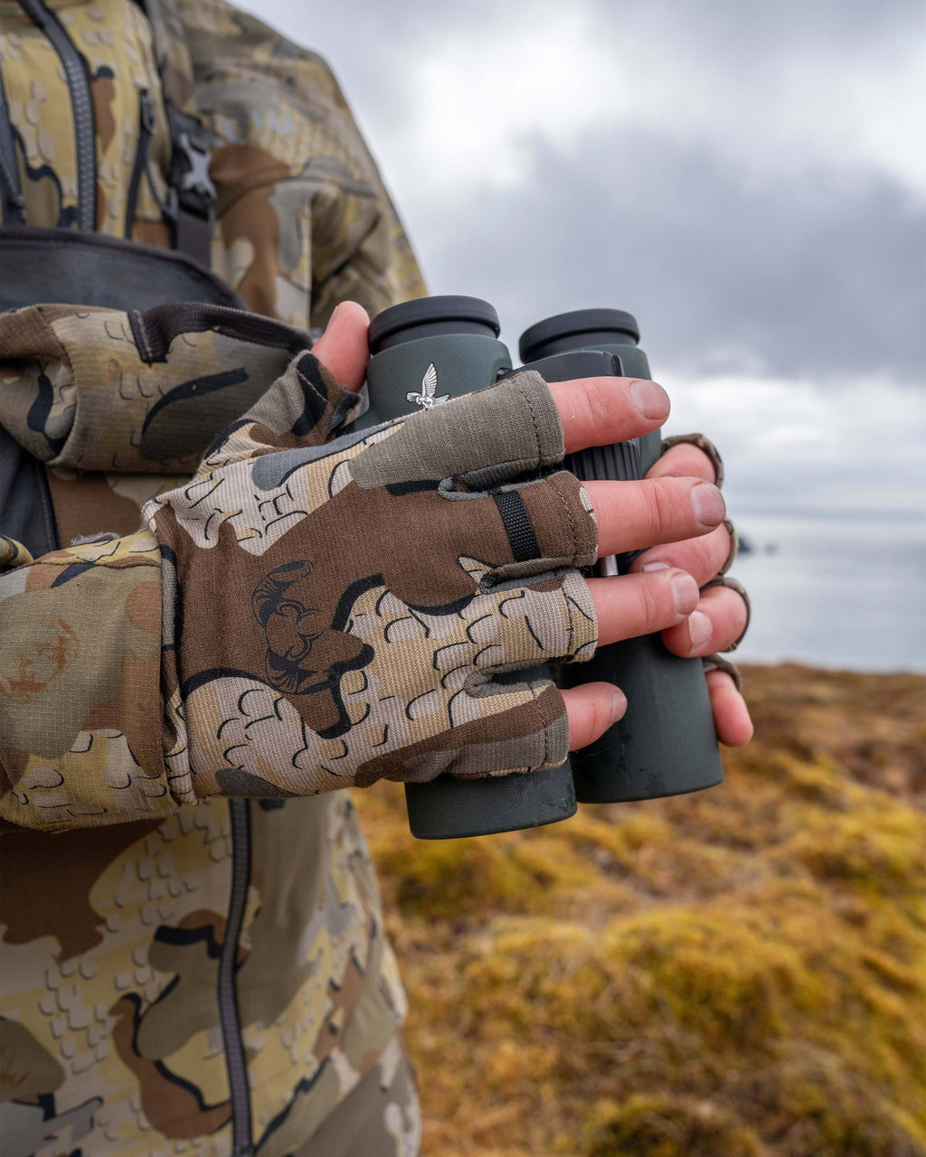 Hunter in an outdoor scene, holding a binocular, wearing ULTRA Merino 210 Fingerless Glove in Valo Camouflage