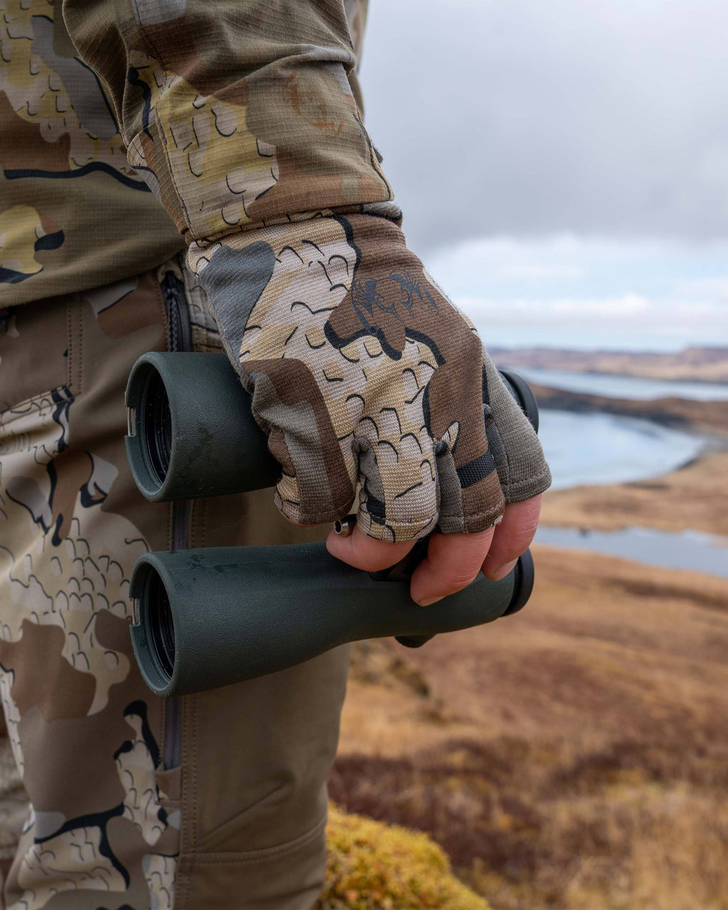 Hunter in an outdoor scene, holding a binocular down on his side, wearing ULTRA Merino 210 Fingerless Glove in Valo Camouflage