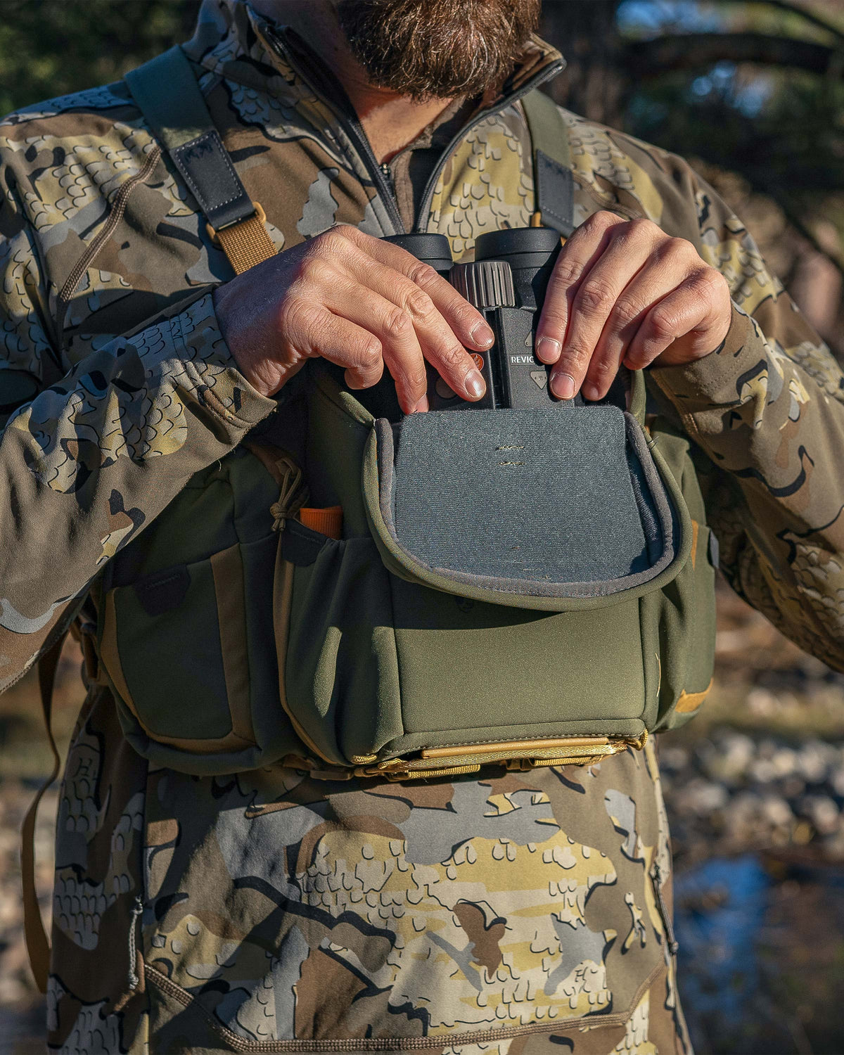 Hunter wearing the PRO Chest Pack in Evergreen, pulling out a binocular from the main compartment, highlighting overlapped forward-folding lid 