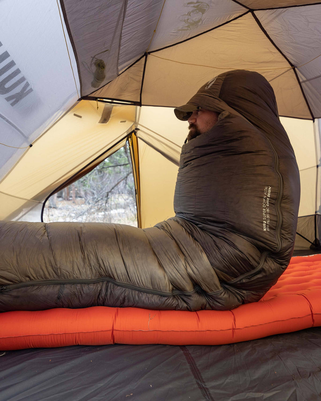 Hunter sitting upright while inside of the fully zipped Super Down Altai 0 degree sleeping bag in Ash Brown with the hood pulled over his head for extra warmth