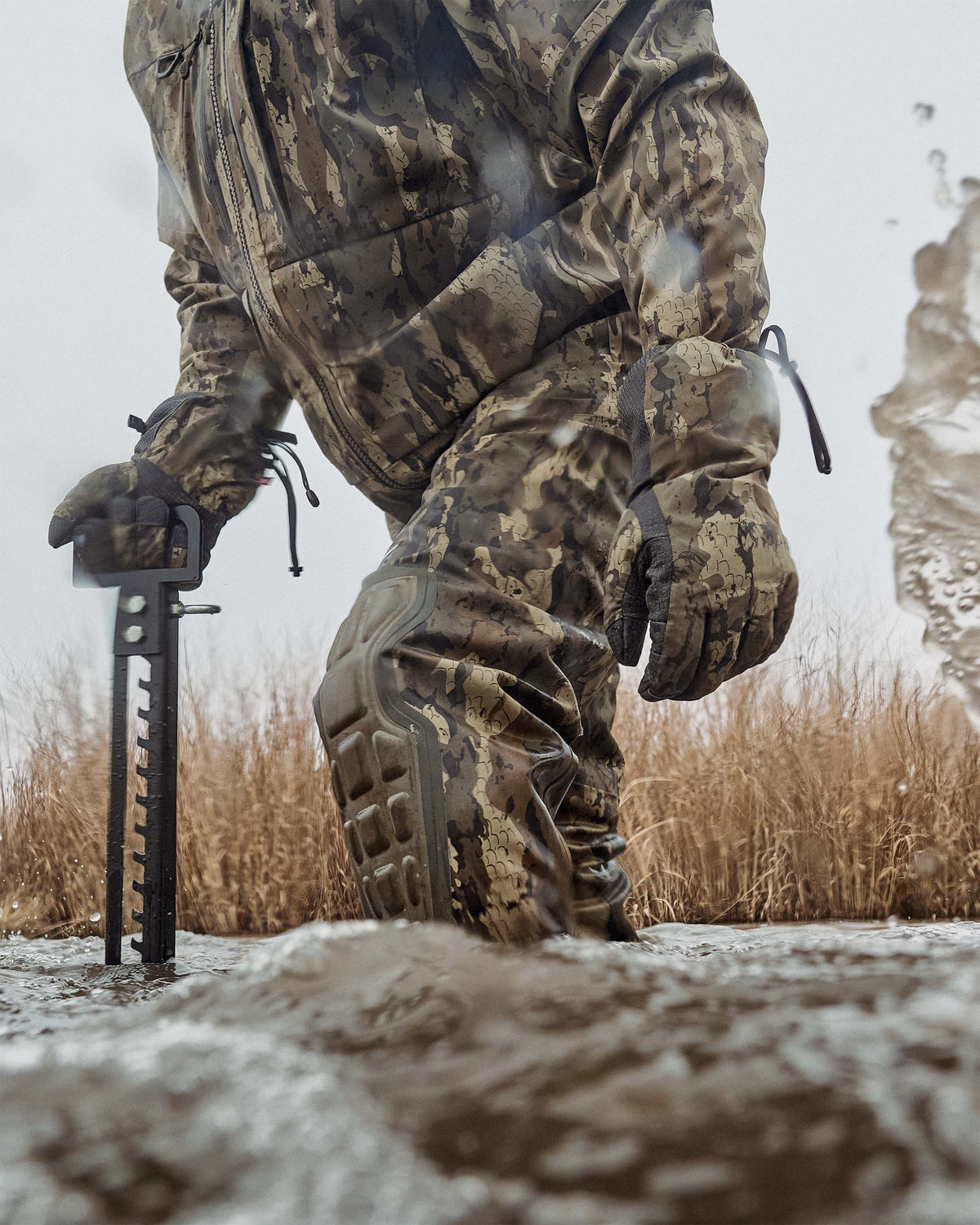 Hunter wearing HD Flex Storm Shell Jacket, KUIU PRO HD Flex Zip Wader and Northstar Glove in Veros camouflage walks through shallow muddy water.  