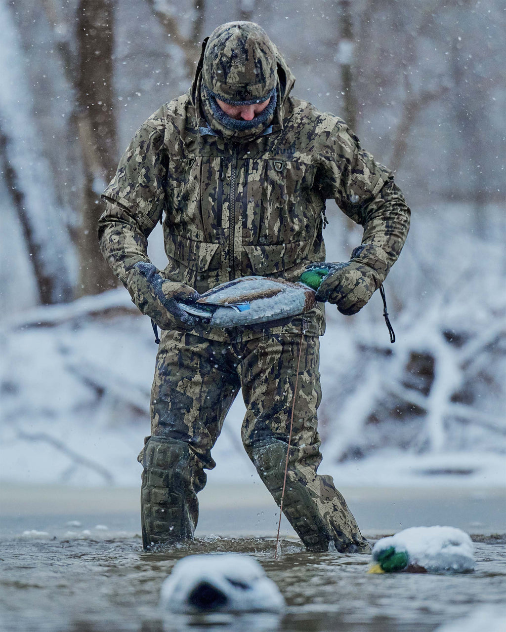 Hunter wearing HD Flex Storm Shell Jacket, KUIU PRO HD Flex Zip Wader and Northstar Glove in Veros camouflage stands in cold, icy, shallow water during a snowstorm, holding a duck decoy. 