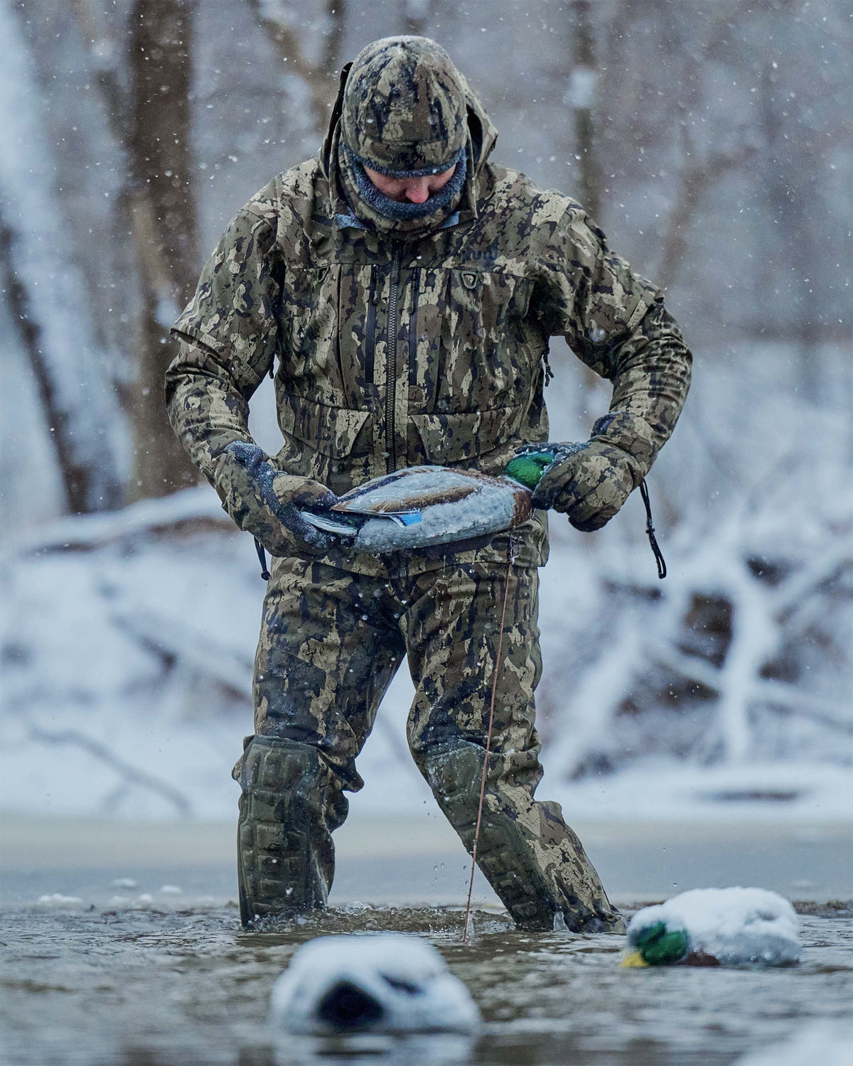 Hunter wearing HD Flex Storm Shell Jacket, KUIU PRO HD Flex Zip Wader and Northstar Glove in Veros camouflage stands in cold, icy, shallow water during a snowstorm, holding a duck decoy. 