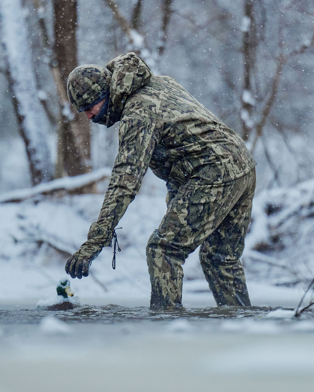 Hunter wearing HD Flex Storm Shell Jacket, KUIU PRO HD Flex Zip Wader and Northstar Glove in Veros camouflage stands in shallow water during a snowy outdoor scene, reaching toward a duck decoy in the water.