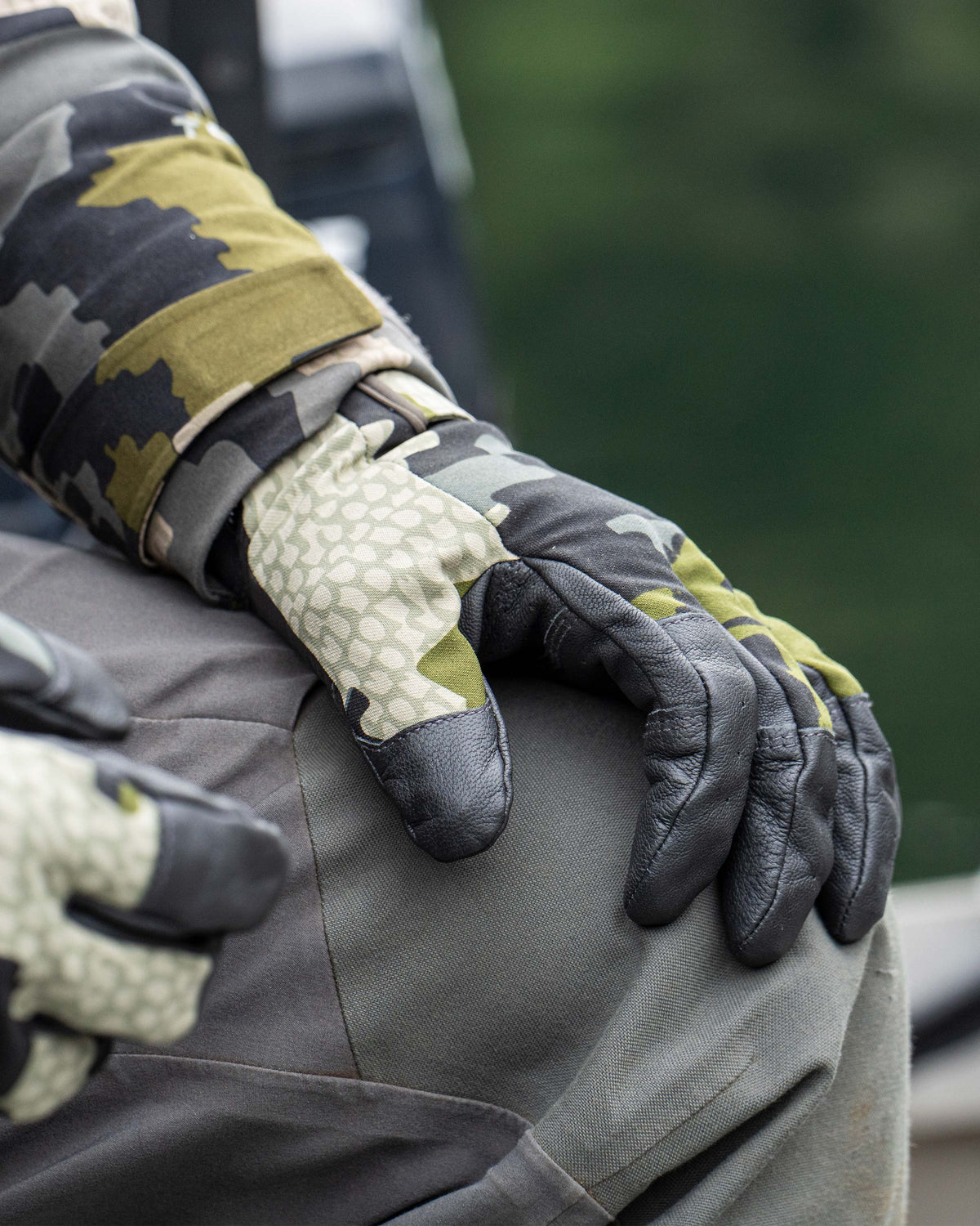 Close-up of hunter left hand on his knee wearing Precision Glove in Verde Camouflage, Highlighting goat skin leather across the grip, breathable and quick-dry material on back of hand and seamless finger patterning at touch points