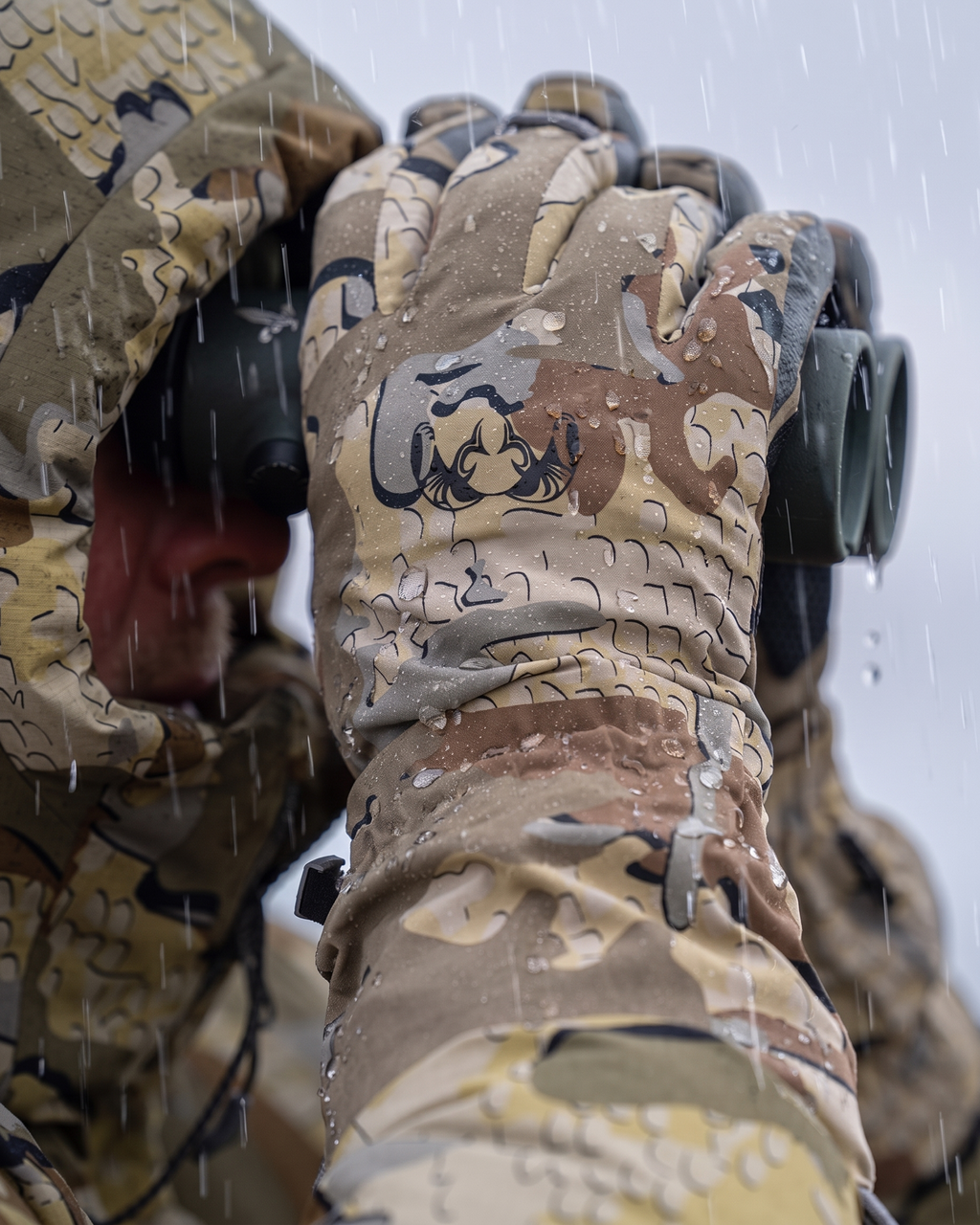 Close-up of a hunter wearing Yukon Hardshell Glove in Valo camouflage matching camouflage clothing, looking through binoculars in the rain.