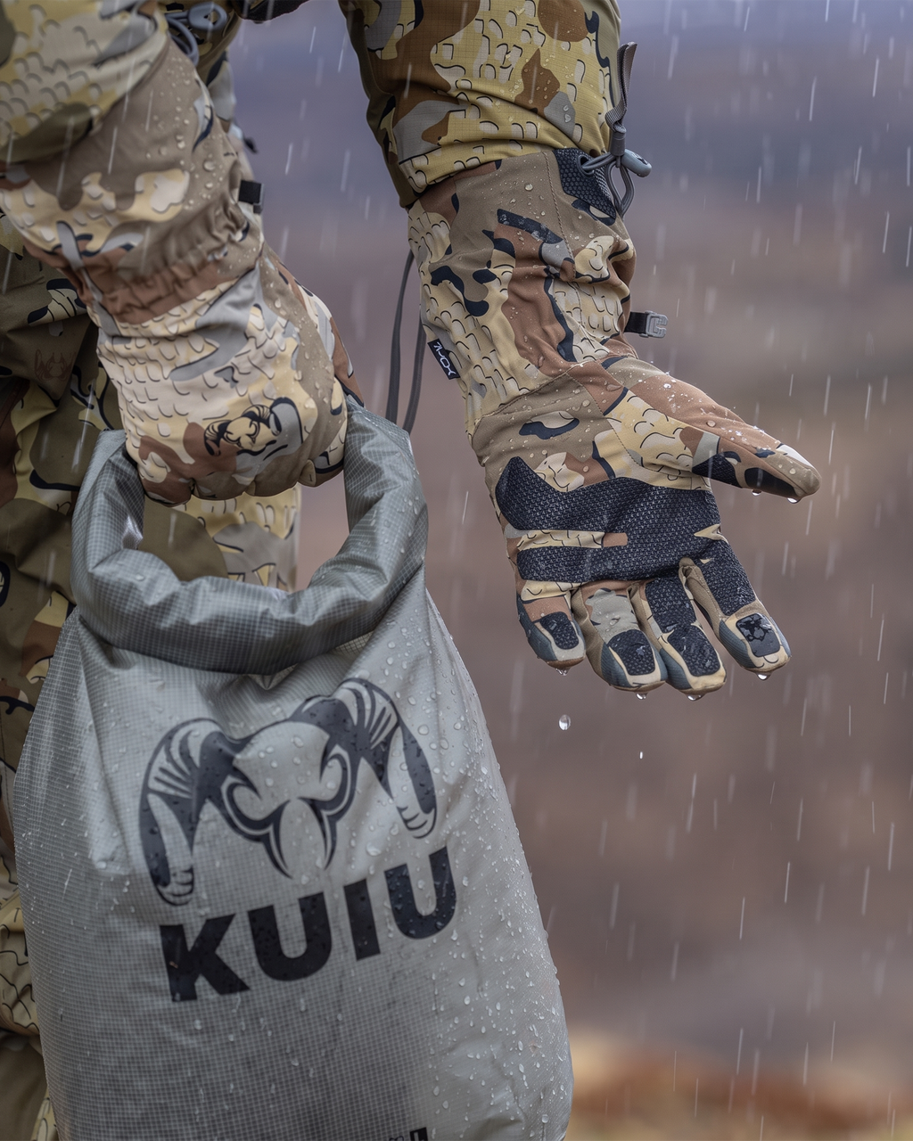 A close-up of a hunter in the rain wearing Yukon Hardshell Glove in Valo camouflage, showing the inner palm, holding a gray KUIU dry bag with a ram logo. 