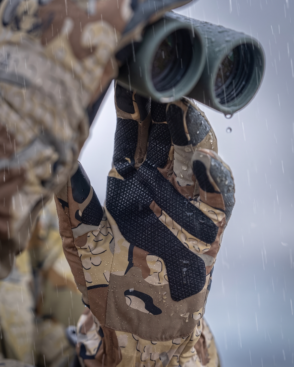 Close-up of a hunter in the rain wearing Yukon Hardshell Glove in Valo camouflage holding a pair of binoculars, with a blurred outdoor background.