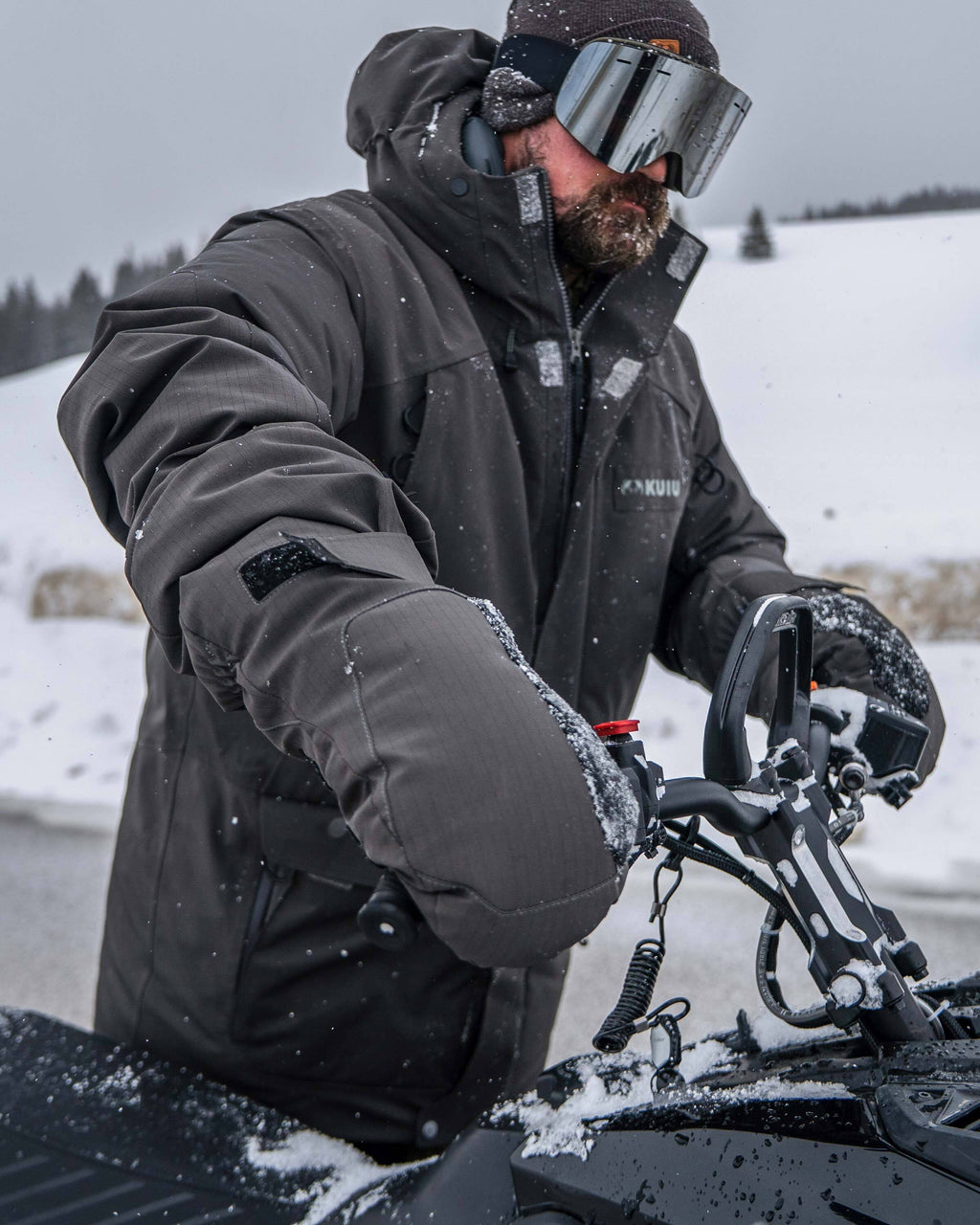Hunter gripping the handlebars of a snowmobile wearing Super Down Arktuk Parka and Super Down Arktuk Mitt in Asphalt Grey