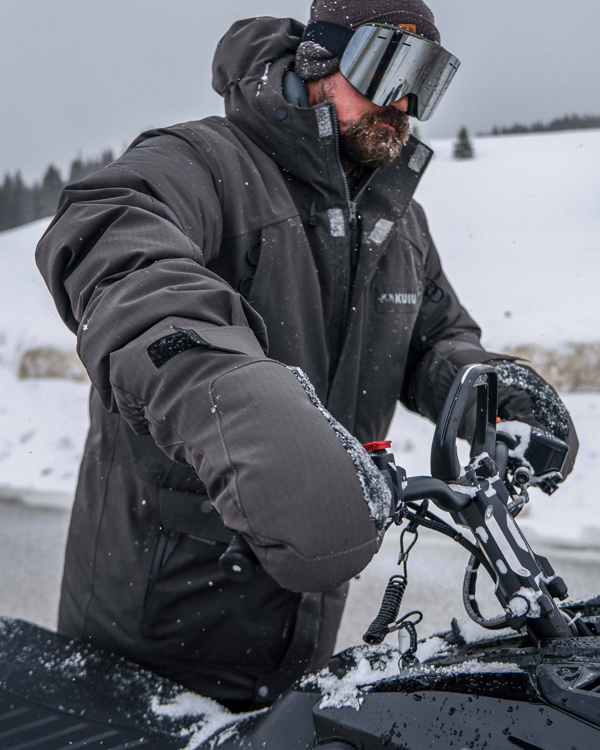 Hunter gripping the handlebars of a snowmobile wearing Super Down Arktuk Parka and Super Down Arktuk Mitt in Asphalt Grey
