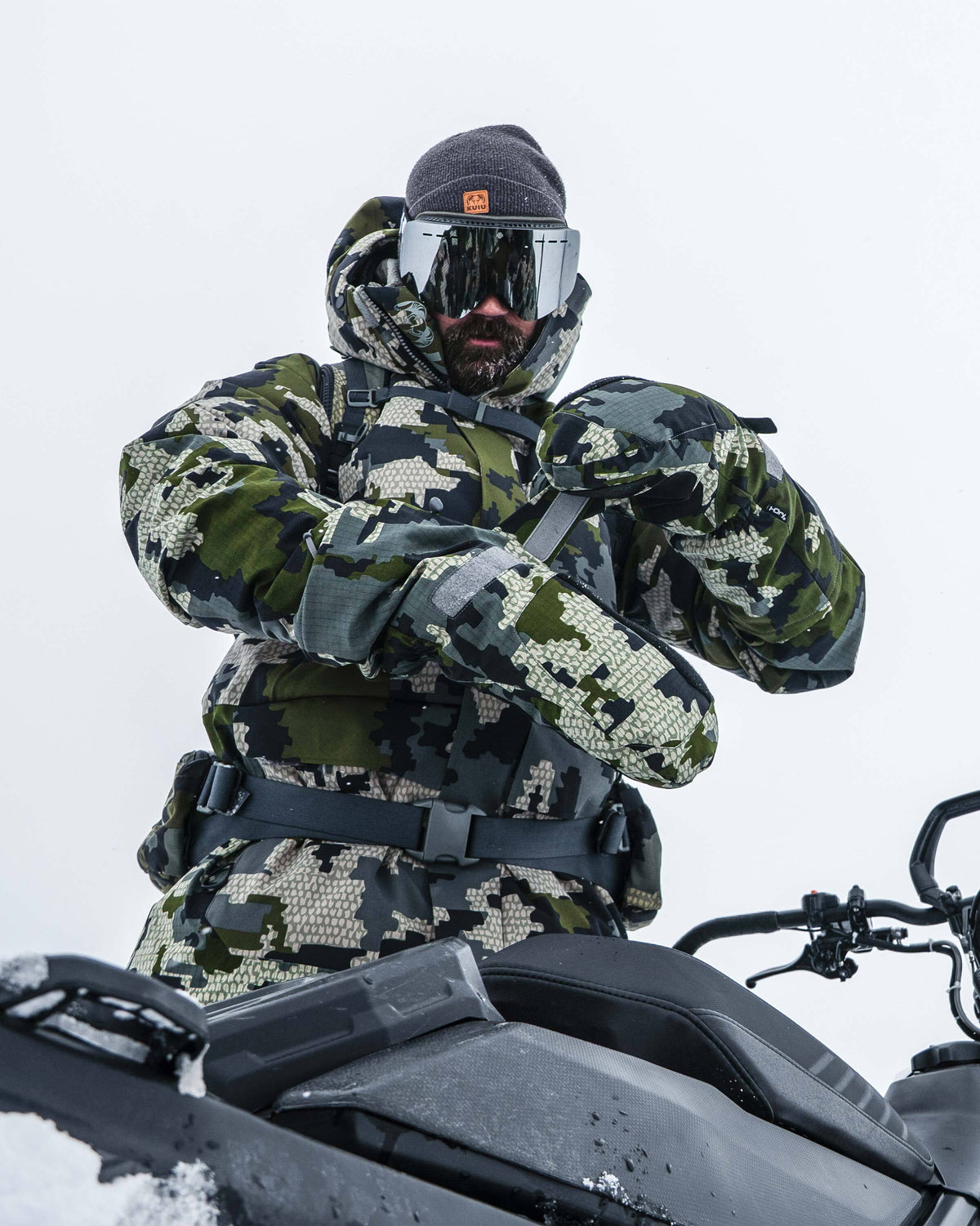 Hunter in a snowy cold climate standing next to a snowmobile, strapping on Super Down Arktuk Mitt in Verde Camouflage