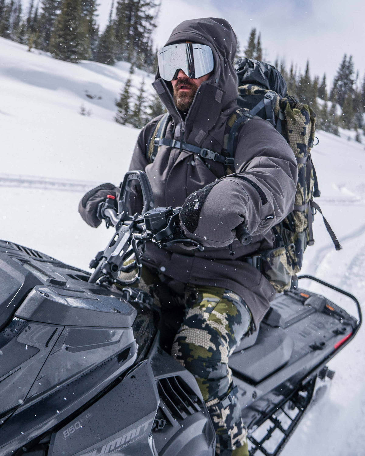 Hunter wearing Super Down Arktuk Parka and Super Down Arktuk Mitt in Asphalt Grey riding a snowmobile through a snowy landscape