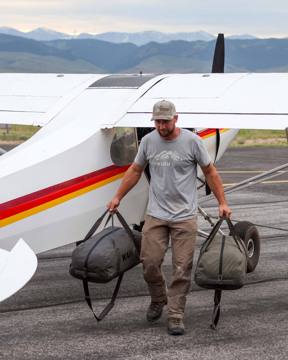 Man unloaded Runway CORDURA® Duffel in Asphalt Grey and Ash Grey from a  Super Cub plane holding one duffel in each hand