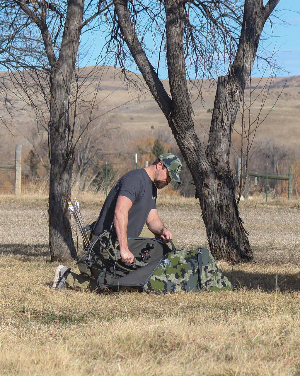 Hunter Kneeling in Open Field Putting Bow Inside of Bow Travel Case in Verde Camouflage