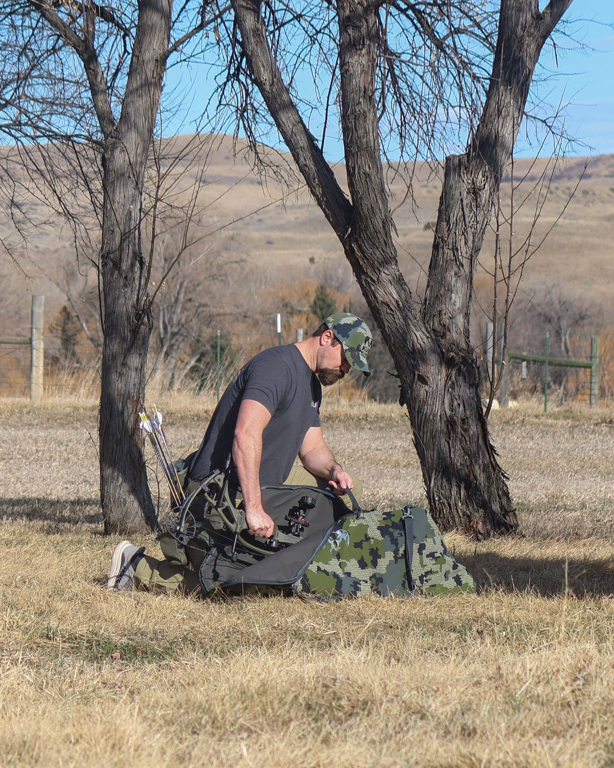Hunter Kneeling in Open Field Putting Bow Inside of Bow Travel Case in Verde Camouflage