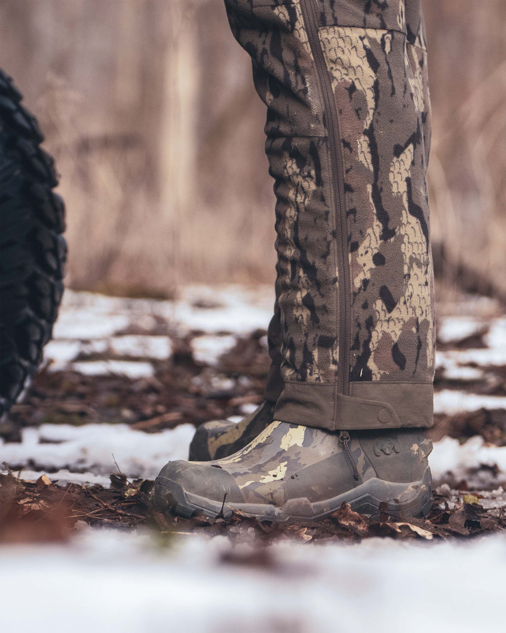 Close-up of a person's lower legs wearing KUIU Mud Boot in Veros camouflage, standing outdoors on a snowy forest ground.