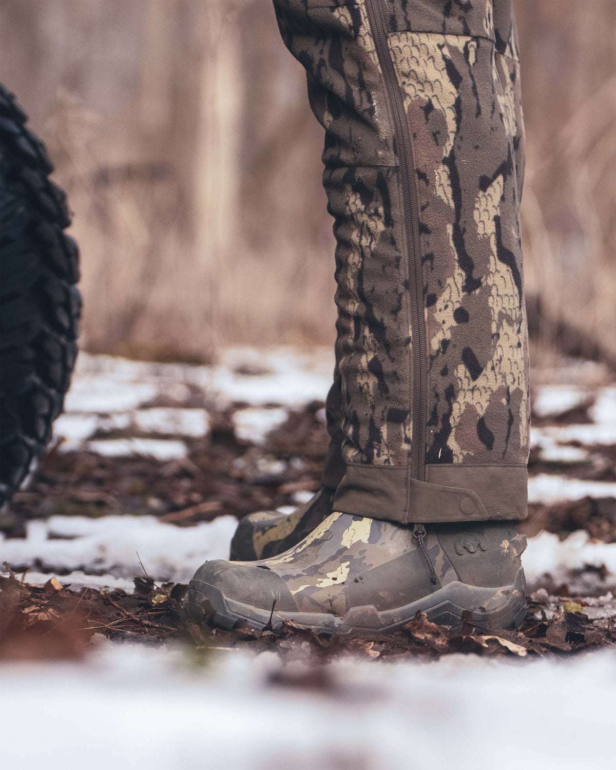 Close-up of a person's lower legs wearing KUIU Mud Boot in Veros camouflage, standing outdoors on a snowy forest ground.