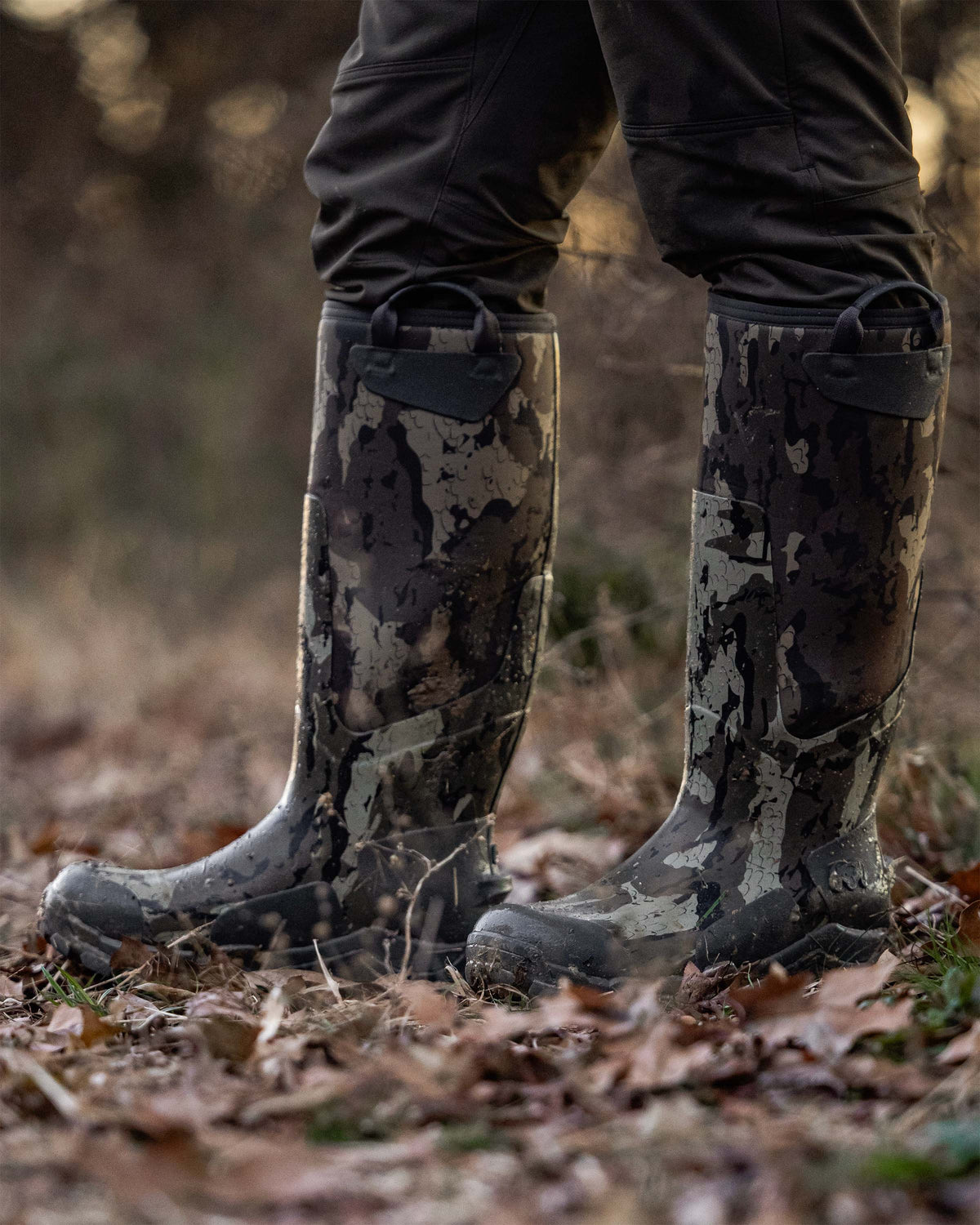 A close-up of a person wearing KUIU Mud Boot in Veros camouflage standing on a forest floor covered in dry leaves.