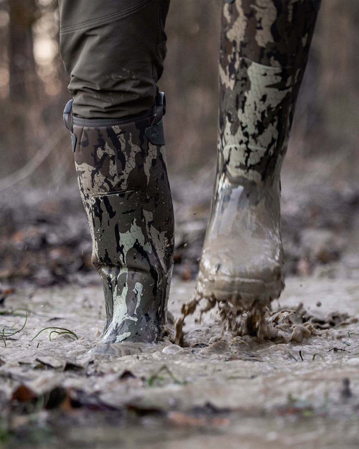 Close-up of a person wearing KUIU Mud Boot in Veros camouflage walking through muddy water outdoors.
