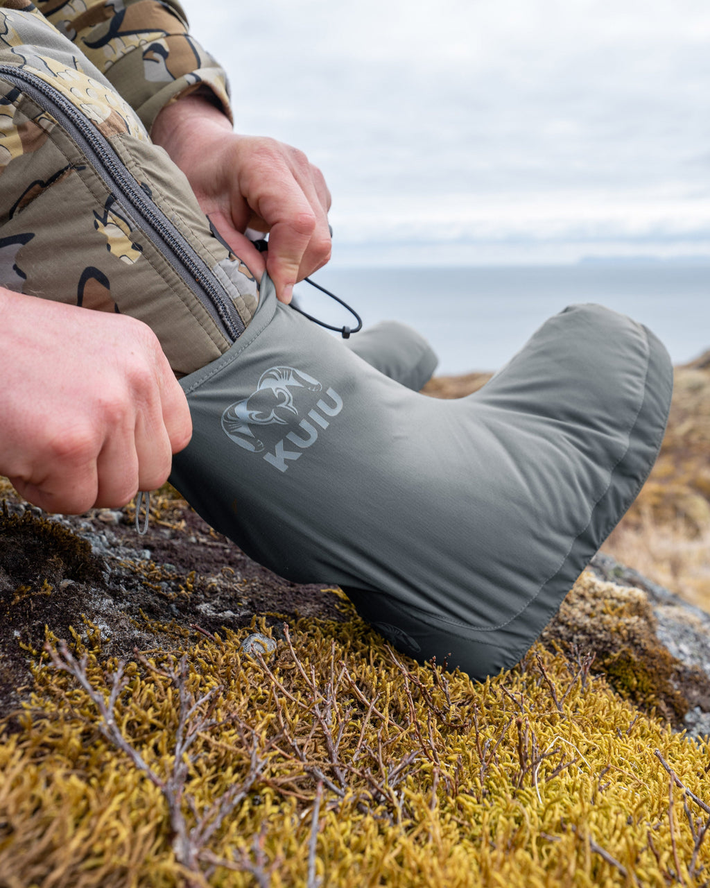 Close-up view of hunter wearing and pulling up the Super Down Burner Bootie in Gunmetal Grey, showing KUIU Ram logo on the bootie
