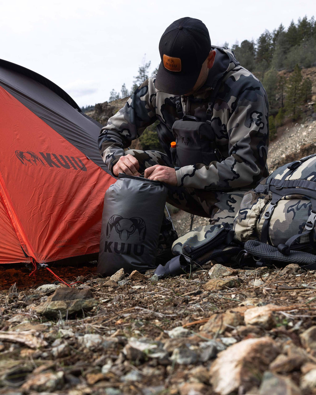 Hunter in Vias Camouflage kneeling to open a closed Stone Grey HD Roll Top Dry Bag
