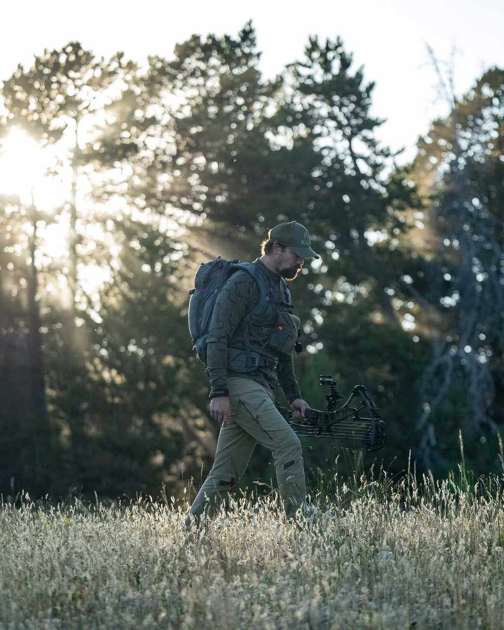 Hunter backpacking through the meadow carrying his bow, wearing Ridgepoint Fleece Henley in Olive Green Tonal Pattern