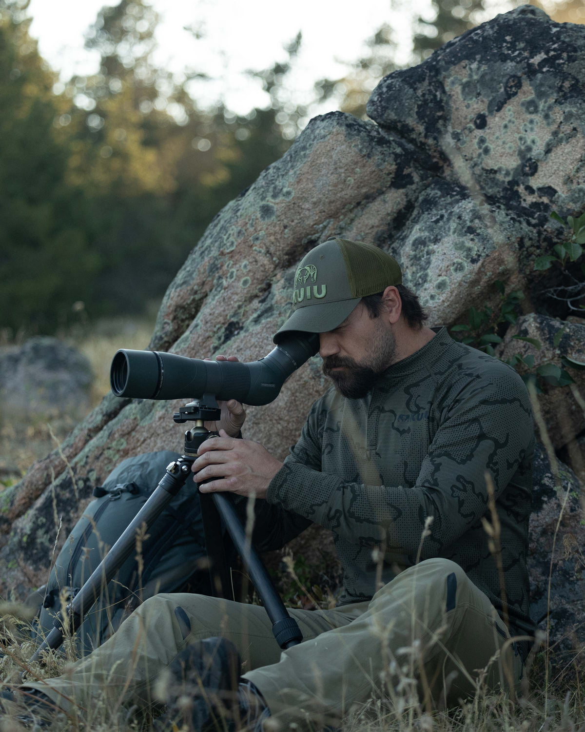 Hunter posted next to a rock, looking through a spotting scope, wearing Ridgepoint Fleece Henley in Olive Green Tonal Pattern