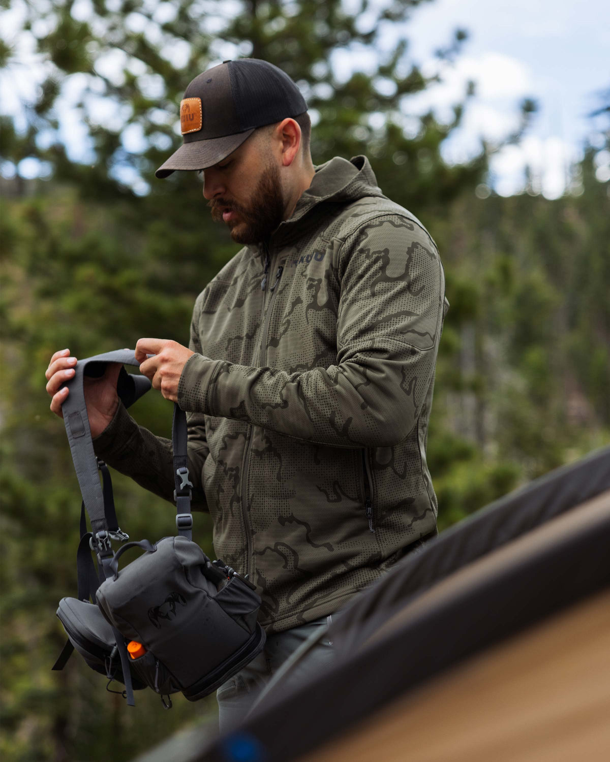 Hunter adjusting his bino harness strap, wearing Ridgepoint Fleece Hoodie in Ash Tonal Brown 