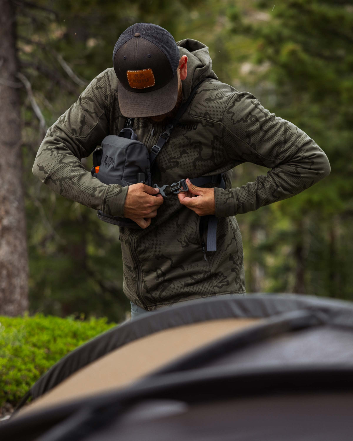 Hunter putting on his bino harness, wearing Ridgepoint Fleece Hoodie in Ash Tonal Brown 
