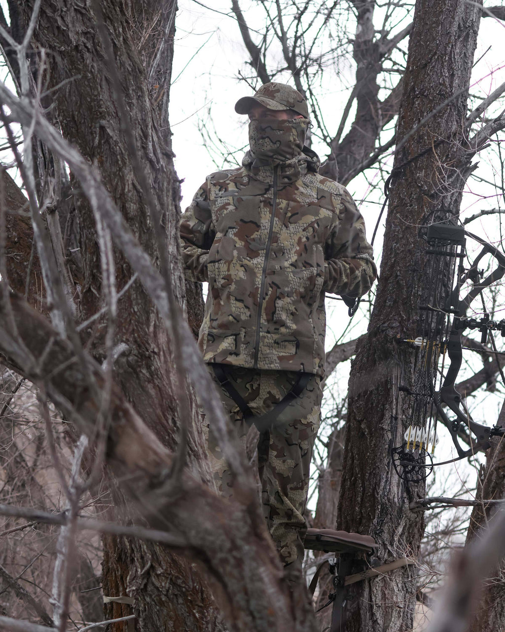Bowhunter standing camouflaged in a tree stand with hands in his pocket, wearing Proximity LT Hooded Jacket in Valo Camouflage