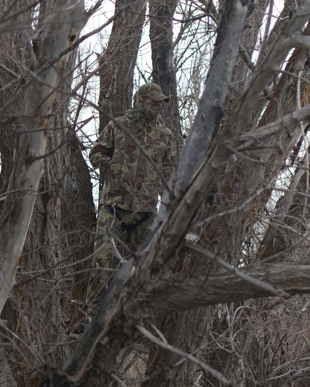 Bowhunter standing camouflaged in a tree stand with hands in his pocket, looking out into the woods, wearing Proximity LT Hooded Jacket in Valo Camouflage