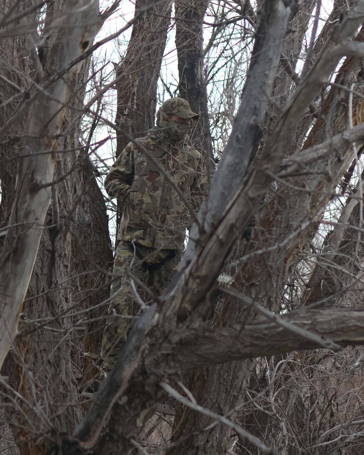 Bowhunter standing camouflaged in a tree stand with hands in his pocket, looking out into the woods, wearing Proximity LT Hooded Jacket in Valo Camouflage